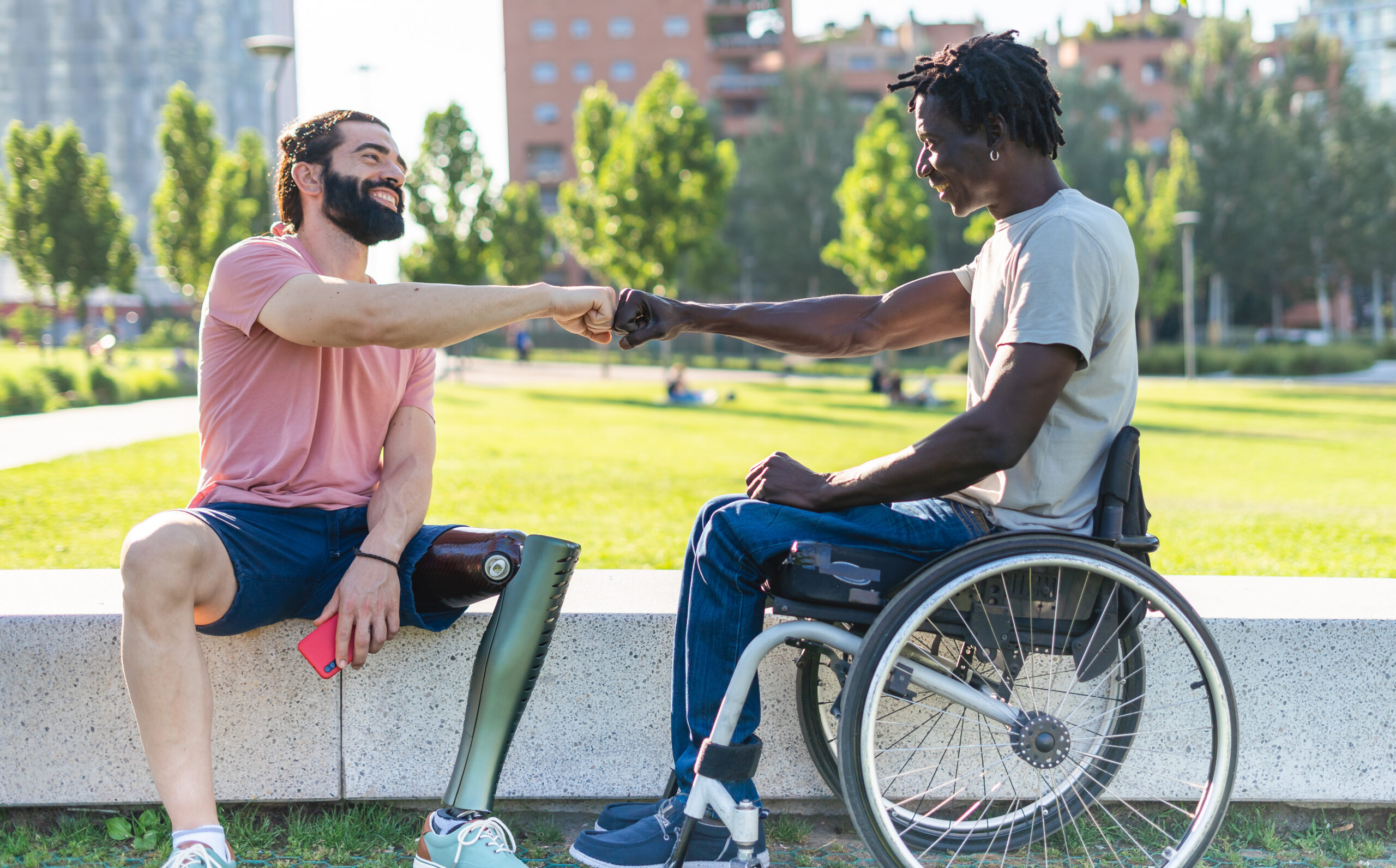 An image of a person who uses a prosthetic leg and a person who uses a wheelchair fist bumping in a park