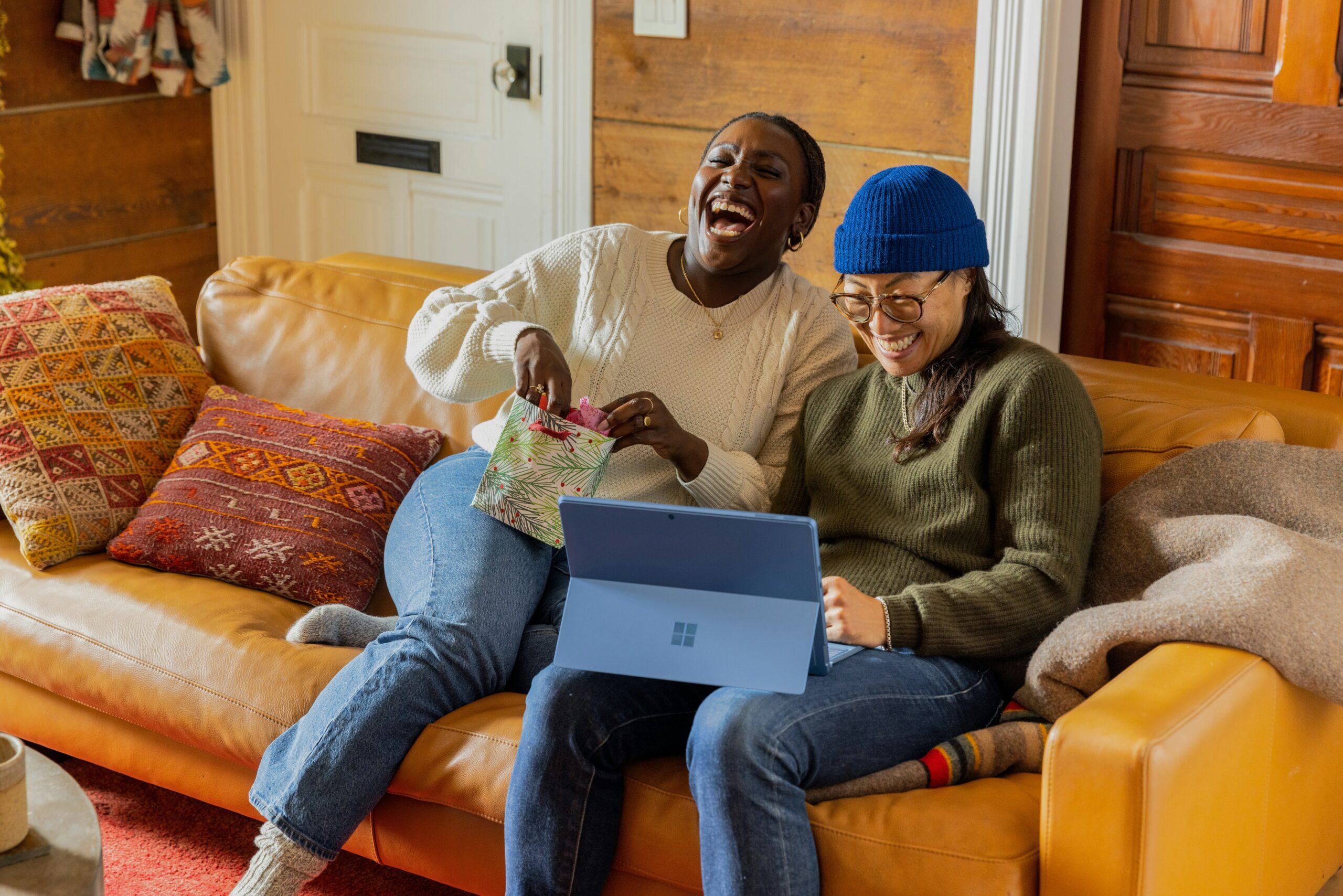 An image of a Black woman, and Asian person sitting together on a couch laughing.